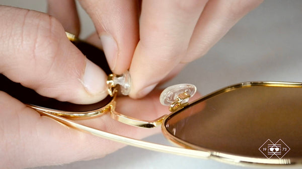 A close-up of a person's hands holding a pair of sunglasses and a small screwdriver. The person is removing the old nose pads from the sunglasses and replacing them with new ones. The new nose pads are white and round.