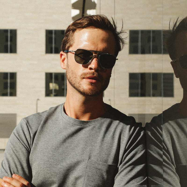 A man in non polarized randolph sunglasses with a reflection of the cityscape on the glass he is leaning on.