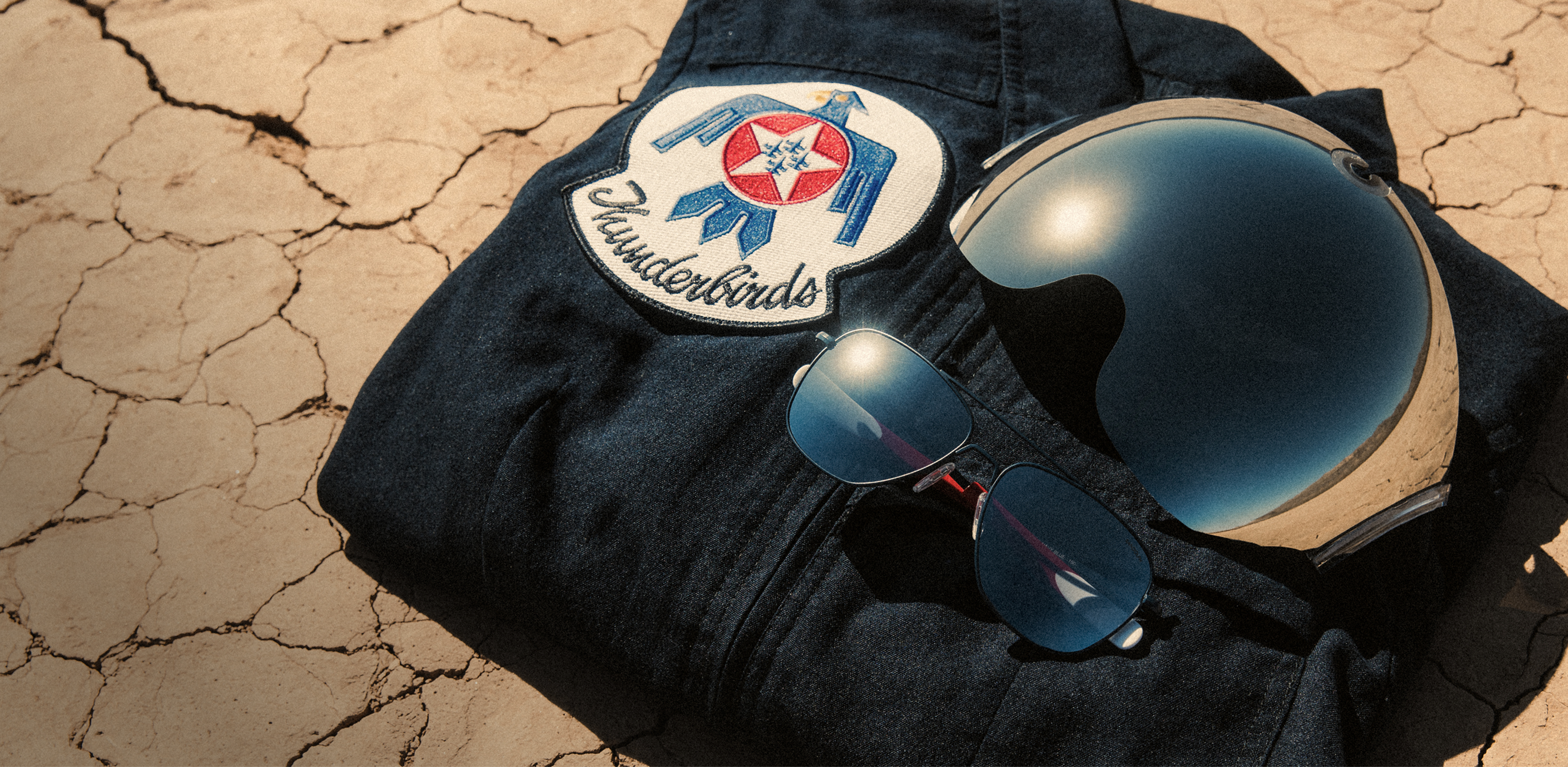 Randolph x Gentex x Thunderbirds Aviator sunglasses with silver mirror lenses beside a Thunderbirds flight helmet and flight suit.